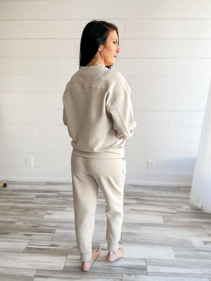 Woman wearing a beige outfit standing in a room with wooden flooring and white walls.